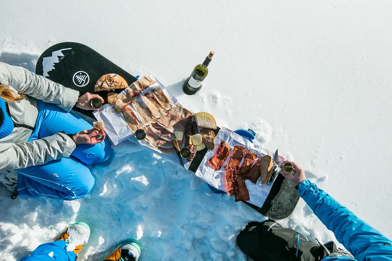 A snowboard is being used as a makeshift table for a spread of food and drink in the snow