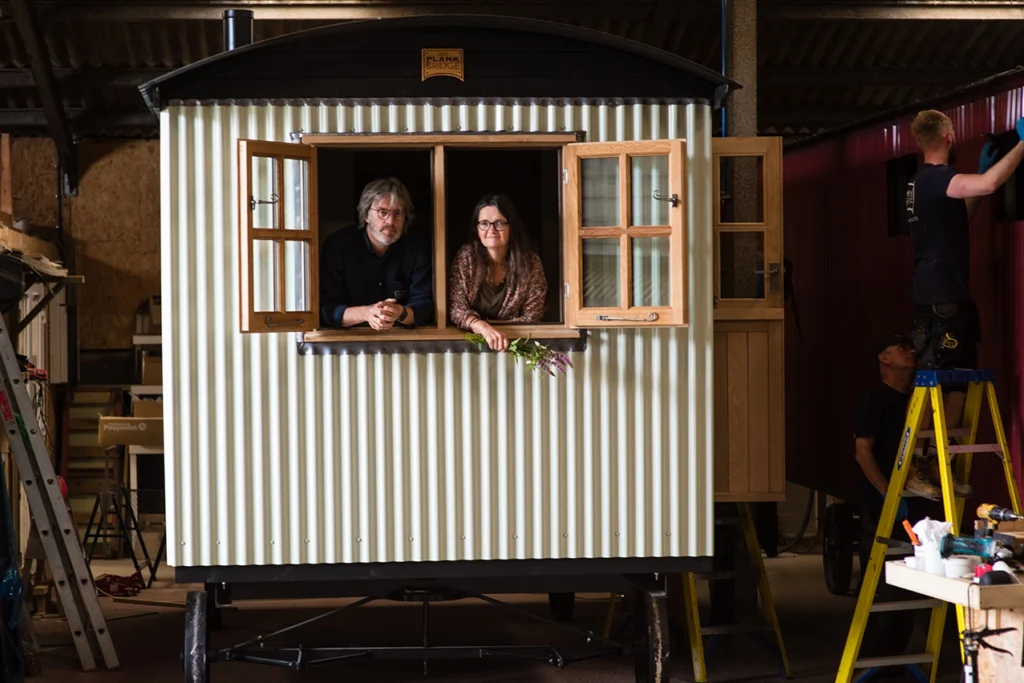 Plankbridge co-founders Richard Lee and Jane Dennison leaning out of a shepherd's hut window. Photographer Katharine Davies