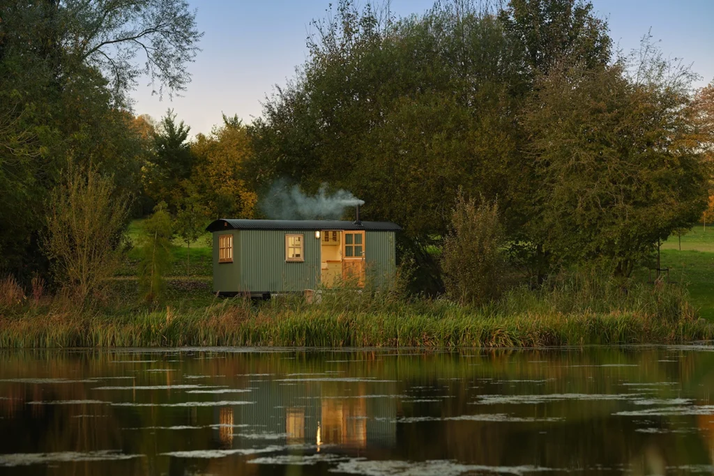 A picturesque Plankbridge shepherd's hut with smoke coming from the chimney sat on the water's edge