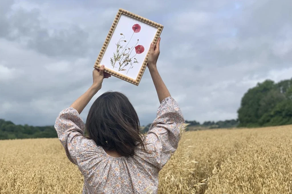 A woman walking through a wheat field wearing a flower print dress holding a framed piece of pressed poppy flowers