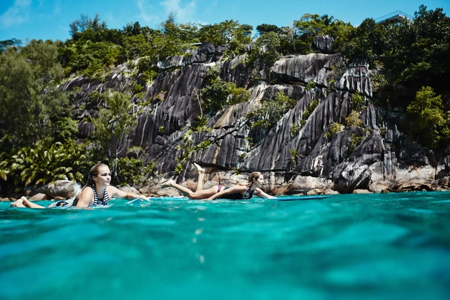 Two women lying on their surf boards, paddling through turquoise water with black cliffs and greenery behind them on the coast.
