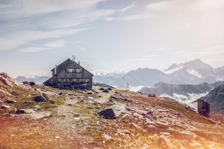 A wooden Alpine lodge sits on a mountain slope with more mountains in the background.