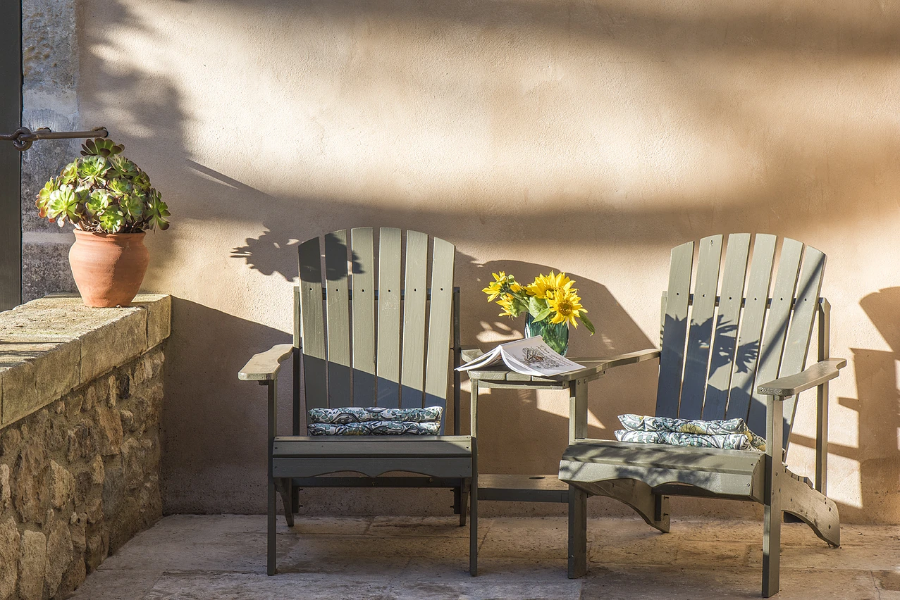 Two wooden slatted deck chairs placed on a terrace at Mas le Gaudre with a small table in between with flowers. The dappled sun hits the wall behind them.