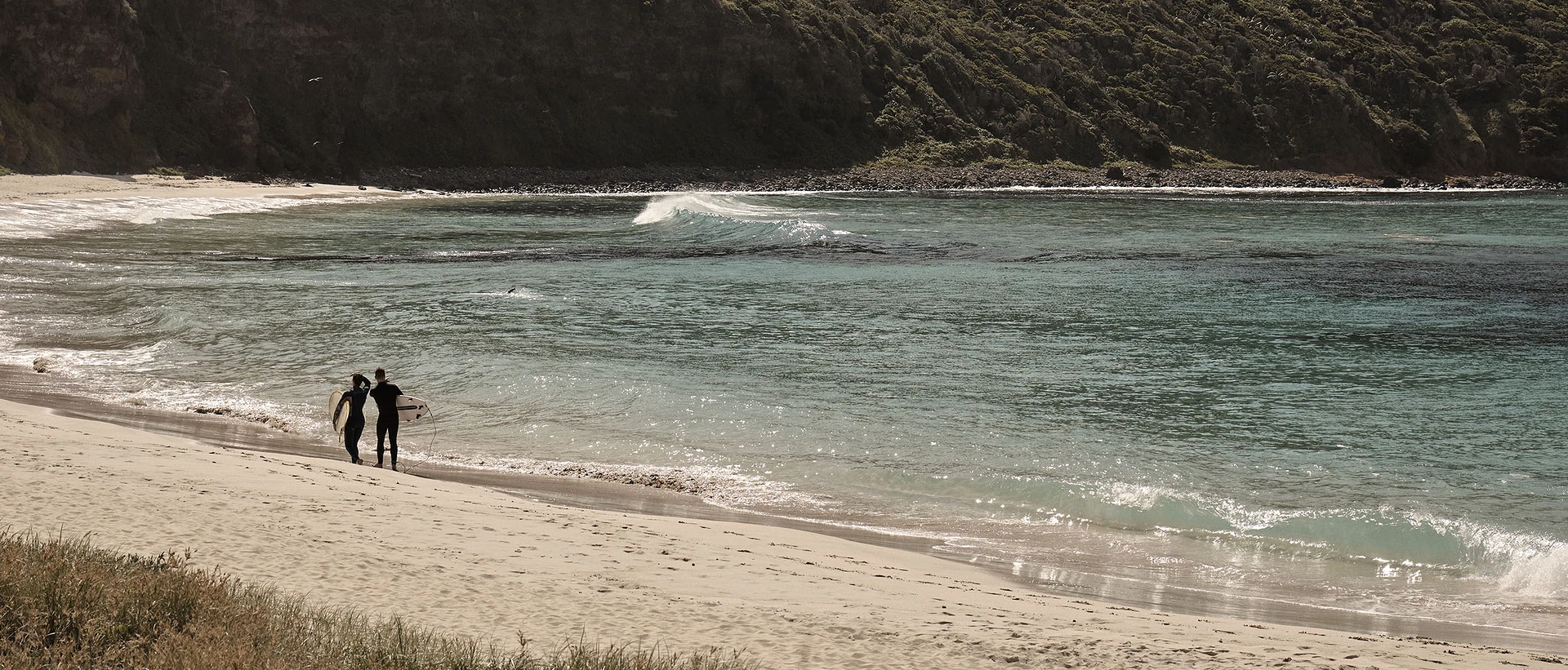 Two surfers carrying boards walk along a sandy beach beside calm turquoise water and rocky cliffs